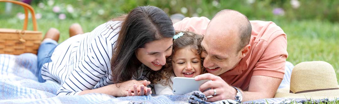 Man, woman, and child laying on a blanket in the grass looking at a smartphone and smiling