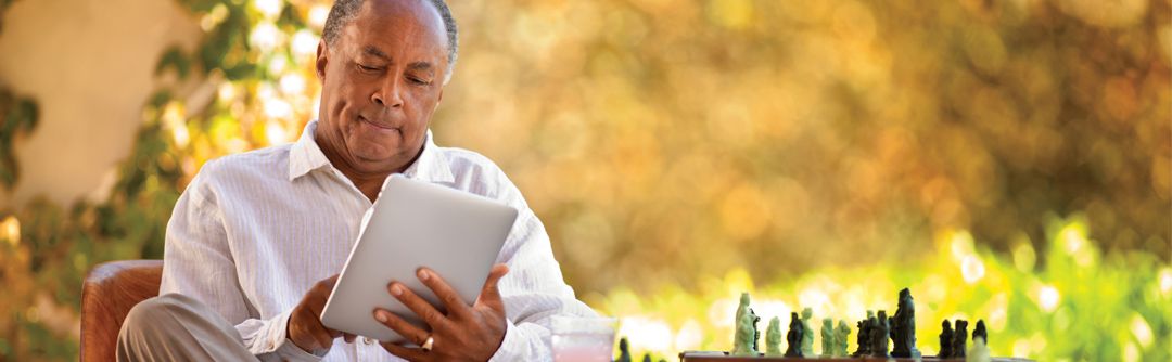 Man sitting outside in a chair next to a chess set; man is looking at and using a tablet