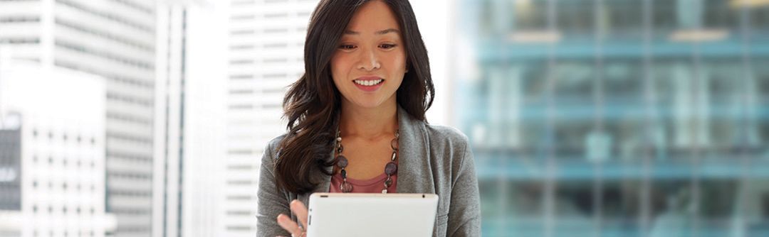 Woman in front of a glass window in business attire smiling and using a tablet