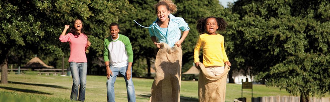 Two adults cheering on two children having a sack race outdoors in the grass