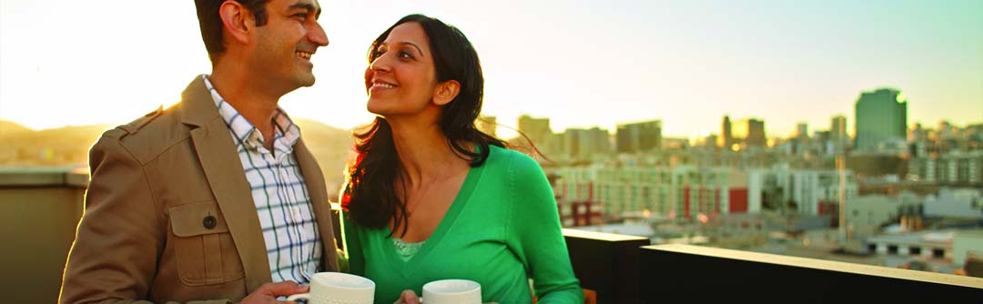 Man and woman smiling at each other, drinking coffee on a rooftop in a city