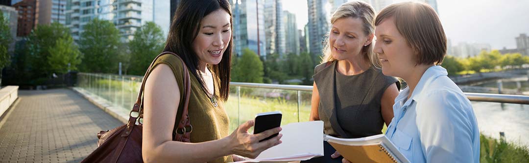 Three women outside on the sidewalk of a city park holding notebooks and documents; one woman is showing the other two something on her cell phone