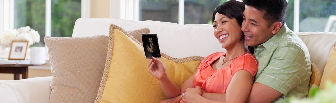 Man and woman sitting on the couch; man has hands around woman's waist and they are looking at a sonogram