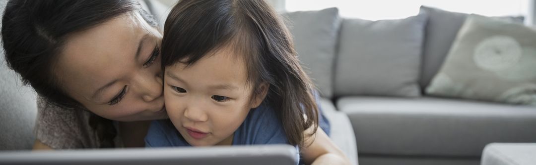 Woman kissing her toddler-aged daughter on the cheek while they both look at a tablet