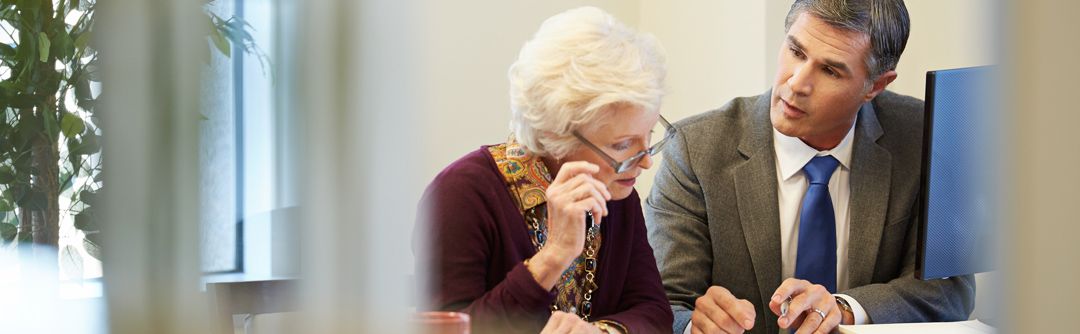 Man and elderly woman sitting at table; woman looking down at documents