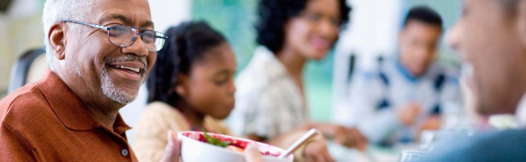 A family sitting down at a meal; one man is in focus smiling and passing a bowl of food to another person and the rest of the family is out of focus