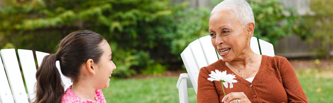 Adult woman and young girl sitting in Adirondack chairs and talking; adult woman has a flower in her hand.