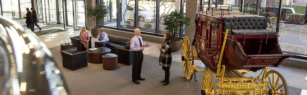 Two people talking in the Wells Fargo Advisors lobby next to the stagecoach. Two more people sitting down on ottomans further away.