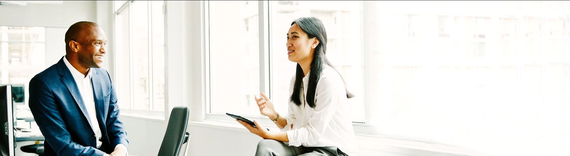 man sitting on desk, woman holding a tablet sitting on chair talking to each other