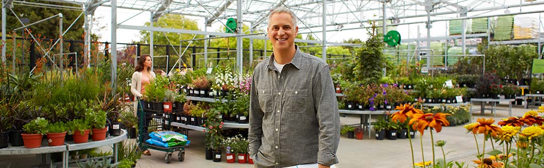 Man standing in a greenhouse smiling at the camera; woman is browsing behind him with a cart full of plants and fertilizer