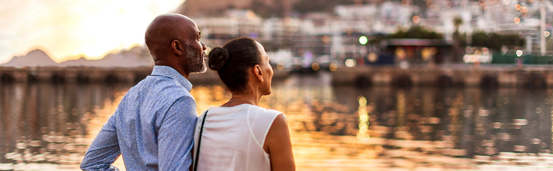 couple standing in front of water way, gazing up towards a community of homes and marina. 