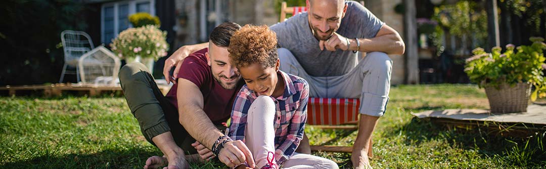 Two fathers playing with their young daughter outside in the grass; one man in sitting in a lawn chair and the other man and child are sitting on the ground