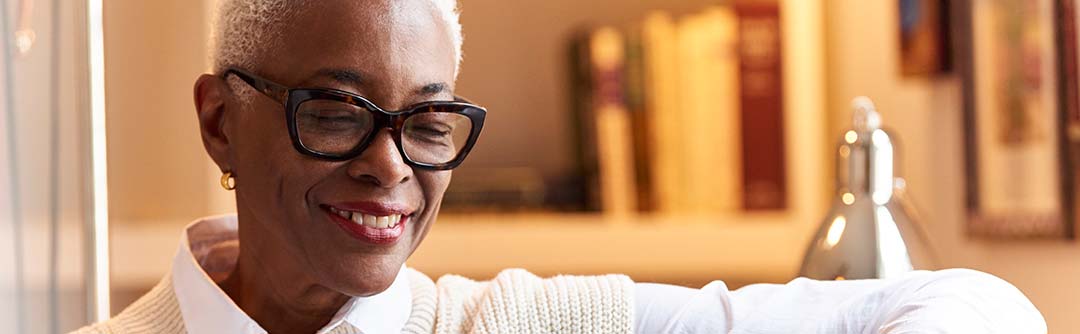 Woman with glasses on sitting in a room with a bookshelf behind her; she is looking down at something and smiling