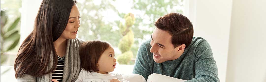 Man, woman, and toddler in the living room of a house; the woman is holding the toddler on the couch and the man leaning over the back of the couch smiling at the toddler