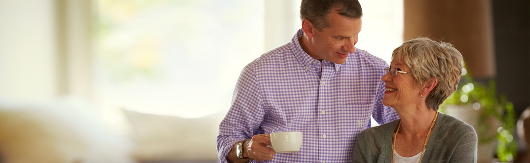 Man and woman in a house; woman is sitting down looking up and smiling at the man who is standing and holding a coffee cup