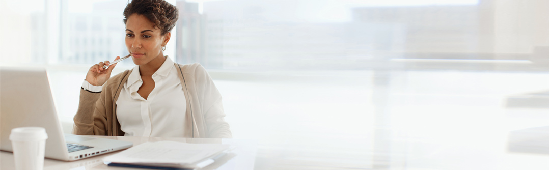 woman sitting in a bright office with large glass windows behind her; she has a pen in her hand and is looking at her laptop with a coffee cup and documents next to the laptop