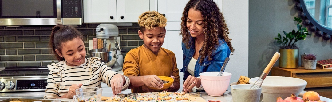 Woman and two children smiling while decorating cookies at a kitchen counter with colorful sprinkles.