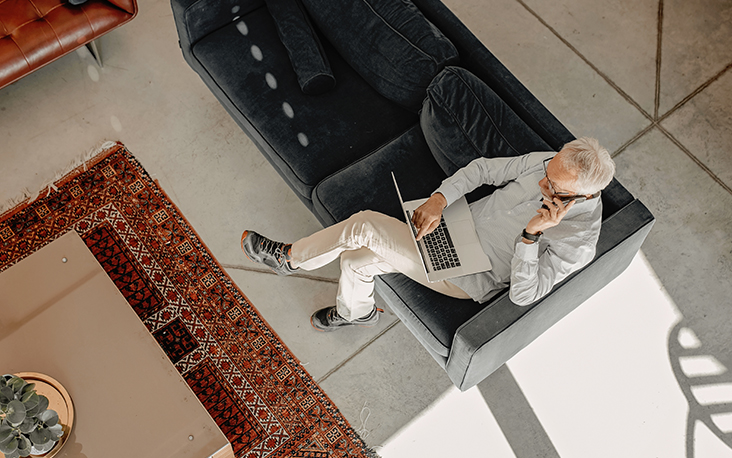 Birds-eye view of mature man sitting on a couch with laptop, and on cell phone validating details with caller