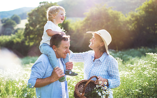 Three people outdoors in a sunlit field, with a child riding on one adult’s shoulders and another adult holding a basket of flowers.