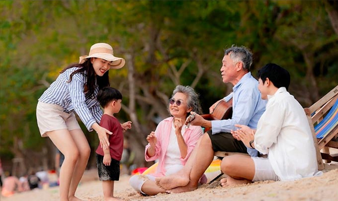 Five people gathered on a beach, including a young child, with several people seated on the sand near lounge chairs.