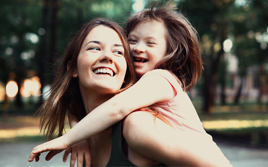 Adult carrying a child piggyback outdoors in a tree-lined park.