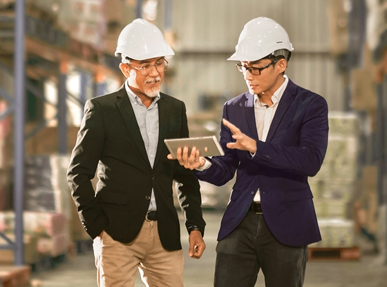 Two men wearing business suits and white safety helmets reviewing a tablet while standing in a warehouse.