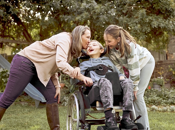 Two women interacting joyfully with a young boy using a wheelchair in a backyard.