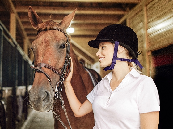 A person wearing a white shirt and black helmet standing next to a brown horse inside a stable.