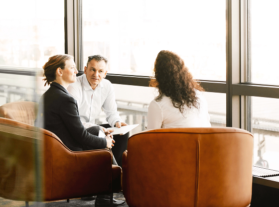 Three people having a discussion while seated in a modern office space near large windows.