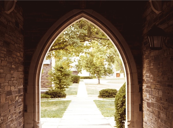 A stone archway framing a view of a tree-lined sidewalk and grassy area outside.
