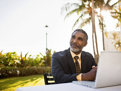 Individual seated outdoors using a laptop on a table in a landscaped area with palm trees and buildings in the background
