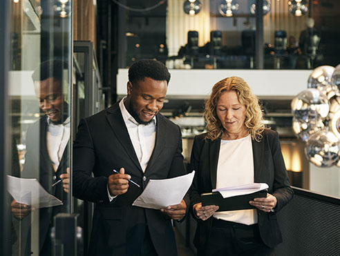 Two business professionals in a modern office reviewing printed documents together near a glass wall