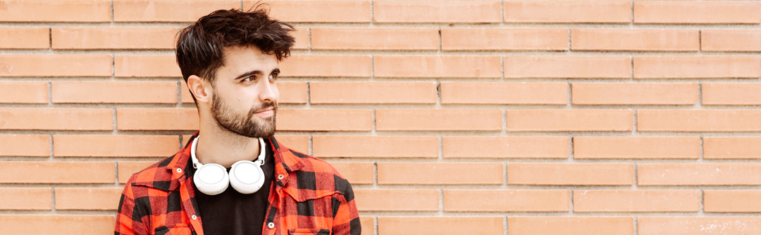 A young man with over-ear headphones around his neck stands in front of a brick wall