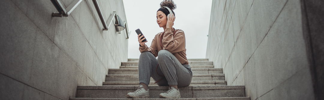 A young woman sits on the steps to a tunnel, looking at her phone and wearing over-ear headphones